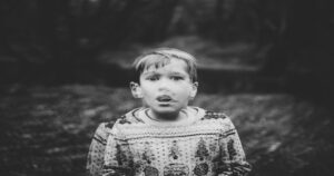 The black-and-white image shows a young boy with a surprised or concerned expression, standing outdoors in a wooded area. Trying to understand Human Trafficking Causes