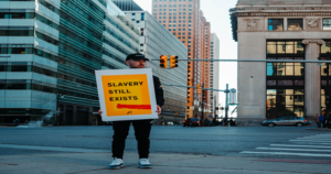 Picture of a man on the corner of a big city. He is holding a sign that say slavery still exits.