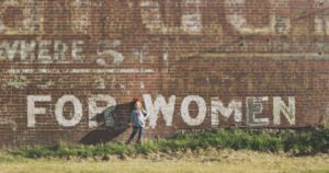 A woman standing in grassy scrub, with her back against a wall. The words "For Women" are panted one the wall. Unveiling the Nexus of Human Trafficking & Technology.